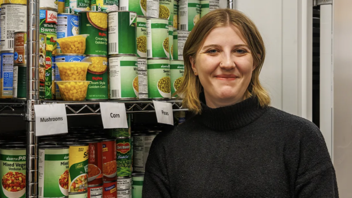 Young woman, Kaitlyn Richards, standing in front of a shelf with canned goods. She has short blonde hair and is smiling while wearing a black sweater.