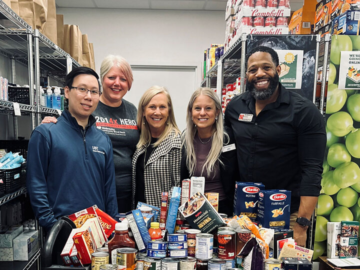 Five individuals stands with a large group of donated food items, inside Husker Pantry.